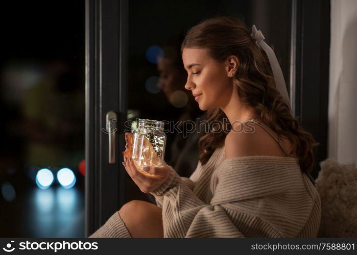 winter, comfort and people concept - young woman in pullover sitting at window with garland lights in glass mason jar mug at home. woman with garland lights in glass mug at home