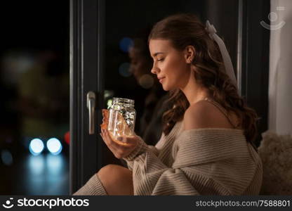 winter, comfort and people concept - young woman in pullover sitting at window with garland lights in glass mason jar mug at home. woman with garland lights in glass mug at home