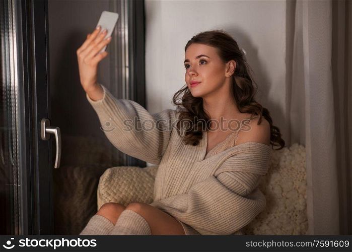 winter, comfort and people concept - young woman in pullover sitting at window at home and taking selfie with smartphone at night. woman taking selfie with smartphone at window