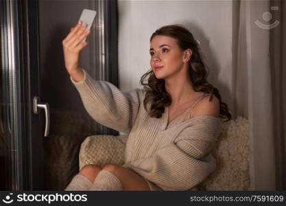 winter, comfort and people concept - young woman in pullover sitting at window at home and taking selfie with smartphone at night. woman taking selfie with smartphone at window