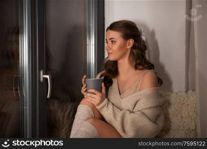 winter, comfort and people concept - young woman in pullover sitting at window with coffee or tea cup at home. woman with coffee or tea cup at window at home