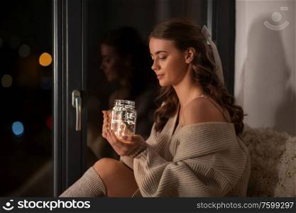 winter, comfort and people concept - young woman in pullover sitting at window with garland lights in glass mason jar mug at home. woman with garland lights in glass mug at home