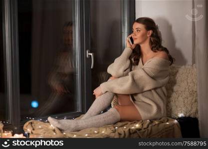 winter, comfort and people concept - young woman in pullover sitting on windowsill at home and calling on smartphone at night. woman sitting on sill and calling on smartphone