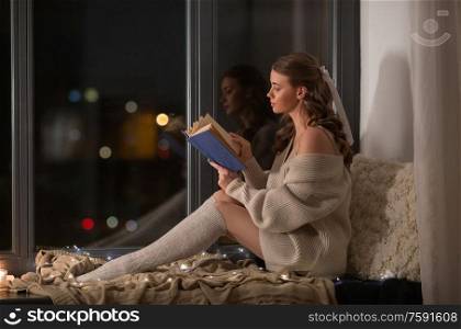 winter, comfort and people concept - young woman in pullover sitting on windowsill and reading book at home. woman reading book sitting on windowsill at home