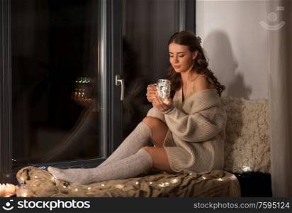 winter, comfort and people concept - young woman in pullover sitting on windowsill with garland lights in glass mason jar mug at home. woman with garland lights in glass mug at home