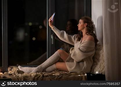 winter, comfort and people concept - young woman in pullover sitting on windowsill at home and taking selfie with smartphone at night. woman taking selfie with smartphone on windowsill