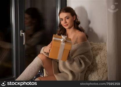 winter, comfort and people concept - young woman in pullover sitting on windowsill with christmas gift at home at night. woman with christmas gift at home at night