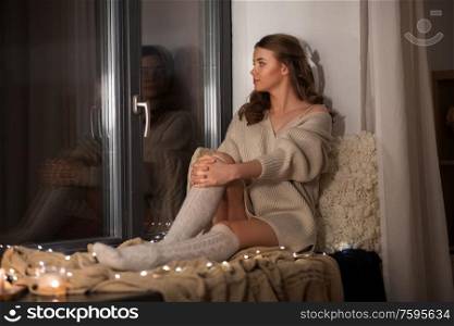 winter, comfort and people concept - young woman in pullover sitting on windowsill at home. woman in pullover sitting on windowsill at home