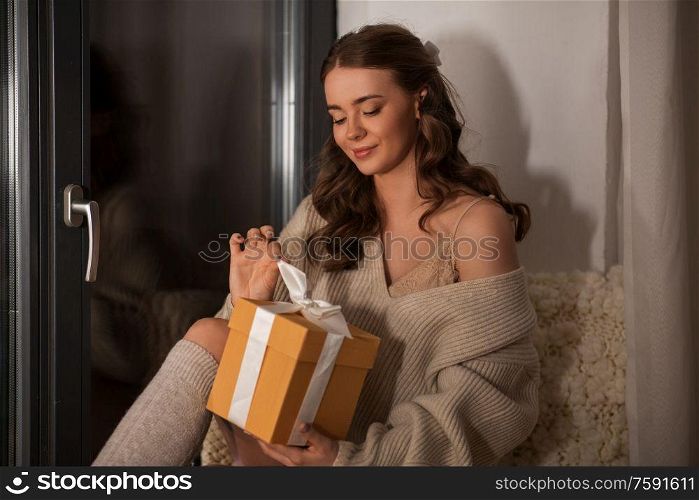 winter, comfort and people concept - young woman in pullover with christmas gift at window at home at night. woman with christmas gift at home at night