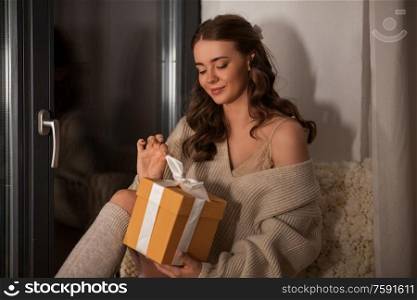 winter, comfort and people concept - young woman in pullover with christmas gift at window at home at night. woman with christmas gift at home at night