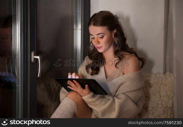 winter, comfort and people concept - young woman in pullover with tablet pc computer sitting at window at home at night. woman with tablet pc sitting at window at home