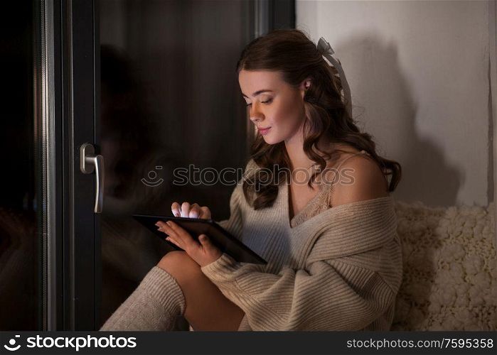 winter, comfort and people concept - young woman in pullover with tablet pc computer sitting at window at home at night. woman with tablet pc sitting at window at home