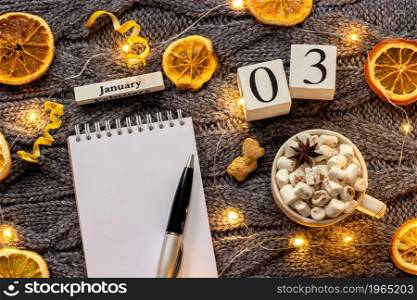 Winter composition. Wooden calendar January 3rd Cup of cocoa with marshmallow, empty open notepad with pen, dried oranges, light garland on grey knitted background. Top view Flat lay Mockup. calendar January 3rd Cup of cocoa and empty open notepad