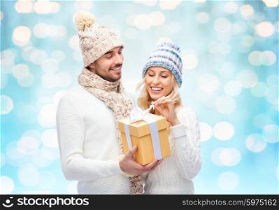 winter, couple, christmas and people concept - smiling man and woman in hats and scarf with gift box over blue holidays lights background