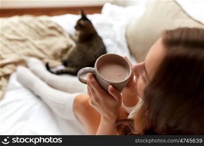 winter, coziness, leisure and people concept - close up of happy young woman with cup of coffee or cacao and cat in bed at home