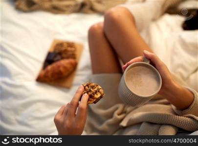 winter, coziness, leisure and people concept - close up of young woman with cup of coffee or cacao and cookie in bed at home