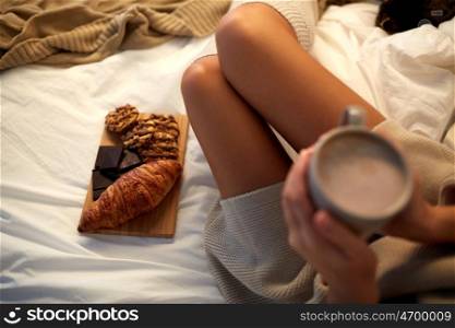 winter, coziness, leisure and people concept - close up of young woman holding cup of coffee or cacao drink with sweets and baking in bed at home