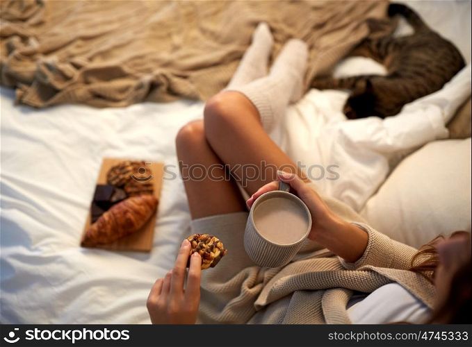 winter, coziness, leisure and people concept - close up of young woman with cup of coffee or cacao and cookie in bed at home
