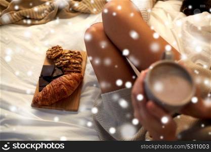 winter, coziness, leisure and people concept - close up of young woman holding cup of coffee or cacao drink with sweets and baking in bed at cosy home over snow. close up of woman with sweets and cocoa in bed