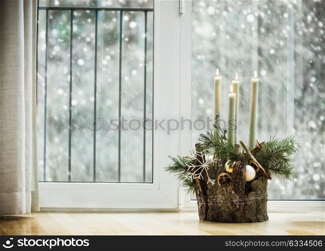 Winter cozy home decoration and festive holiday atmosphere with burning candles, fir branches and snowflakes in living room at window with snowfall. Decorated Advent wreath