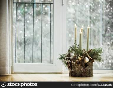 Winter cozy home decoration and festive holiday atmosphere with burning candles, fir branches and snowflakes in living room at window with snowfall. Decorated Advent wreath