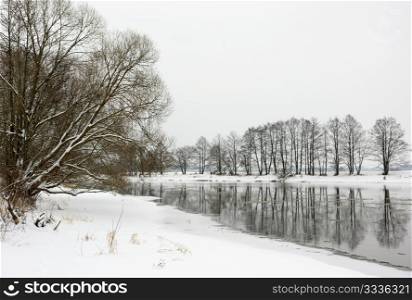 Winter day, snow-covered riverbank, ice, water and trees.