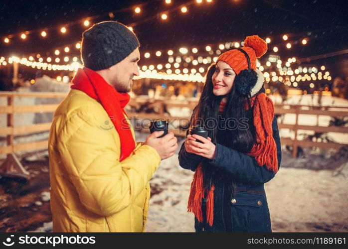 Winter evening, love couple drinks coffee outdoors, holiday illumination on background. Man and woman having romantic meeting on city street with lights. Winter evening, love couple drinks coffee outdoors