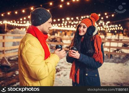 Winter evening, love couple drinks coffee outdoors, holiday illumination on background. Man and woman having romantic meeting on city street with lights. Winter evening, love couple drinks coffee outdoors