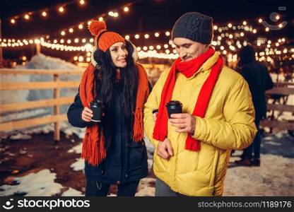 Winter evening, love couple walks with coffee outdoors, holiday illumination on background. Man and woman having romantic meeting on city street with lights. Winter evening, couple walks with coffee outdoors