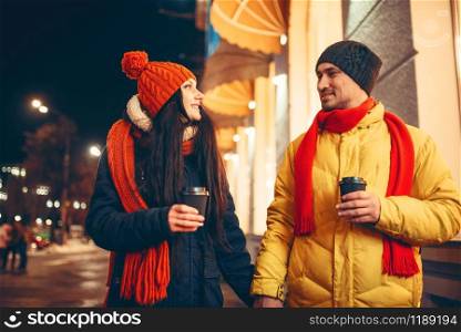 Winter evening, love couple with coffee walking down the street. Man and woman having romantic meeting. Love couple with coffee walking down the street