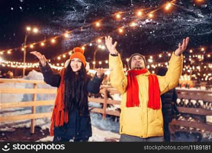 Winter evening, playful love couple throw snow up. Man and woman having romantic meeting on city street with lights