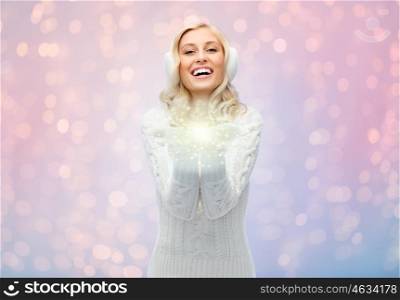 winter, fashion, christmas and people concept - smiling young woman in earmuffs and sweater holding something on her empty palms over rose quartz and serenity lights background