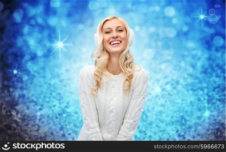 winter, fashion, christmas and people concept - smiling young woman in earmuffs and sweater over blue glitter or lights background