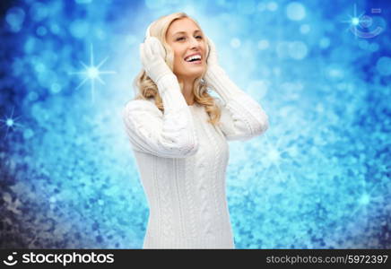 winter, fashion, christmas and people concept - smiling young woman in earmuffs and sweater over blue glitter or lights background