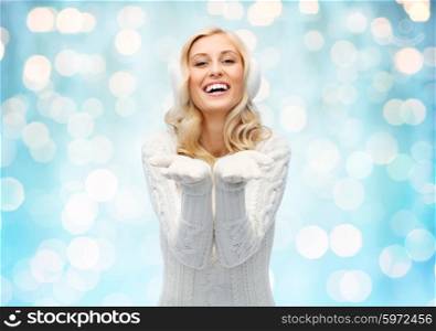 winter, fashion, christmas and people concept - smiling young woman in earmuffs and sweater holding something on her empty palms over blue holidays lights background