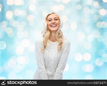 winter, fashion, christmas and people concept - smiling young woman in earmuffs and sweater over blue holidays lights background