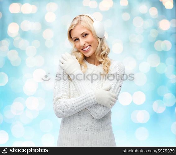 winter, fashion, christmas and people concept - smiling young woman in earmuffs and sweater over blue holidays lights background