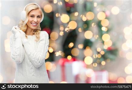 winter, fashion, christmas and people concept - smiling young woman in earmuffs and sweater over holidays lights background