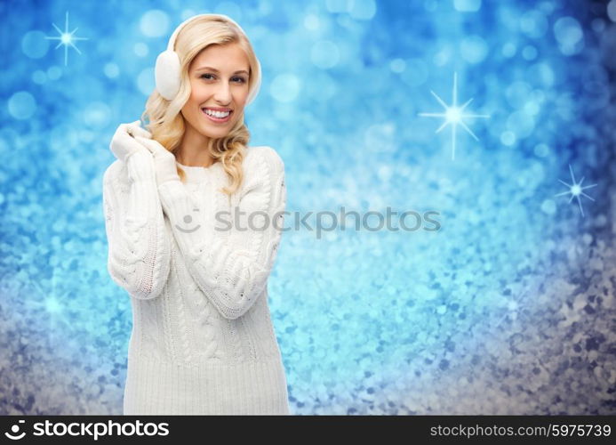 winter, fashion, christmas and people concept - smiling young woman in earmuffs and sweater over blue glitter or holidays lights background