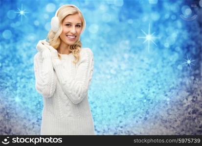 winter, fashion, christmas and people concept - smiling young woman in earmuffs and sweater over blue glitter or holidays lights background