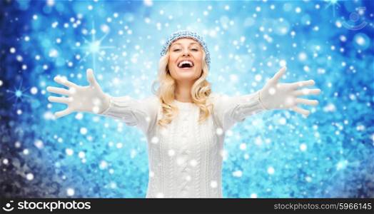 winter, fashion, christmas and people concept - smiling young woman in hat, sweater and gloves over blue lights and snow background