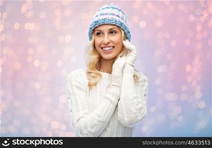 winter, fashion, christmas and people concept - smiling young woman in winter hat, sweater and gloves over rose quartz and serenity lights background