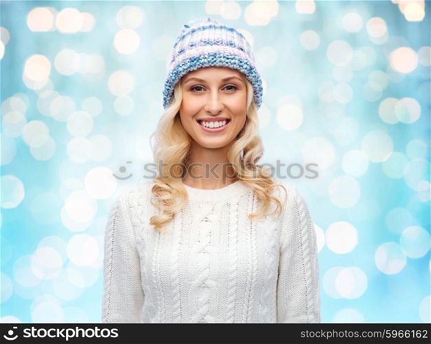 winter, fashion, christmas and people concept - smiling young woman in winter hat, sweater over blue holidays lights background