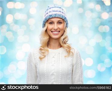 winter, fashion, christmas and people concept - smiling young woman in winter hat, sweater over blue holidays lights background