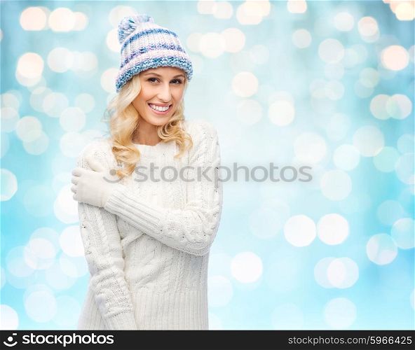 winter, fashion, christmas and people concept - smiling young woman in winter hat, sweater and gloves over blue holidays lights background