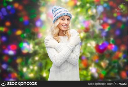 winter, fashion, christmas and people concept - smiling young woman in winter hat, sweater and gloves over holidays lights background