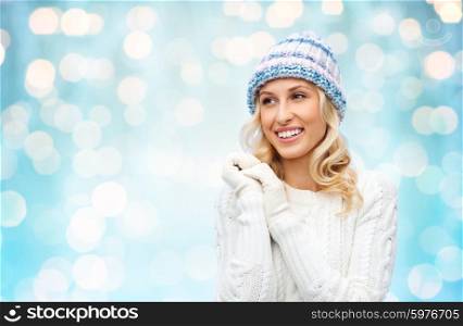 winter, fashion, christmas and people concept - smiling young woman in winter hat, sweater and gloves over blue holidays lights background
