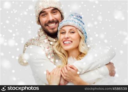 winter, fashion, couple, christmas and people concept - smiling man and woman in hats and scarf hugging over snow background