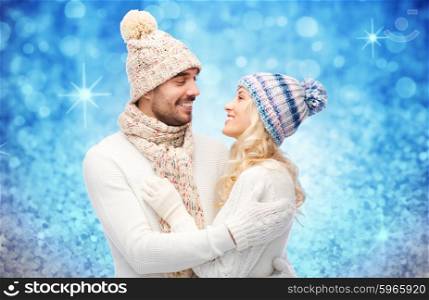 winter, fashion, couple, christmas and people concept - smiling man and woman in hats and scarf hugging over blue glitter and holidays lights background