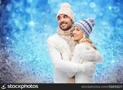 winter, fashion, couple, christmas and people concept - smiling man and woman in hats and scarf hugging over blue glitter or lights background